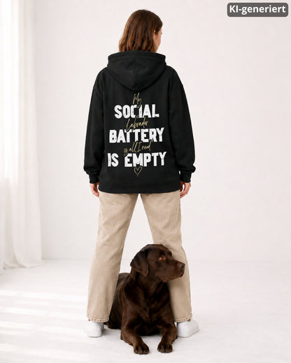 KI-generiertes Modelbild: Frau steht mit dem Rücken zur Kamera und trägt einen schwarzen Oversized Hoodie mit großem Backprint-Motiv „Social Battery is empty“, ein brauner Labrador liegt zwischen ihren Beinen und schaut zur Seite.