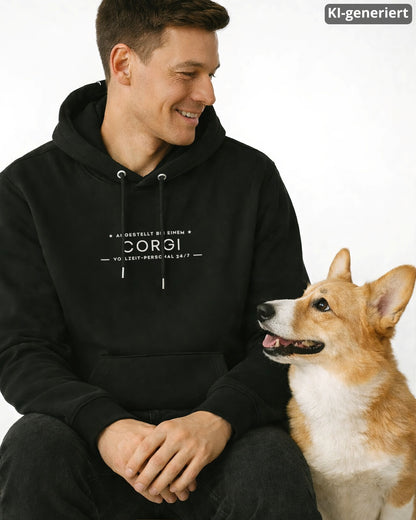 KI-generiertes Modelbild: Mann trägt einen schwarzen Organic Hoodie mit Brustprint-Motiv „Angestellt bei einem Corgi – Vollzeit-Personal 24/7“, daneben sitzt ein Corgi und schaut zu ihm hoch.