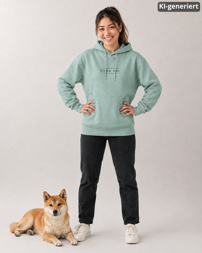 KI-generiertes Modelbild: Frau trägt einen Organic Hoodie in Aloe mit Brustprint-Motiv „Angestellt bei einem Shiba Inu – Vollzeit-Personal 24/7“, daneben liegt ein Shiba Inu im Studio.
