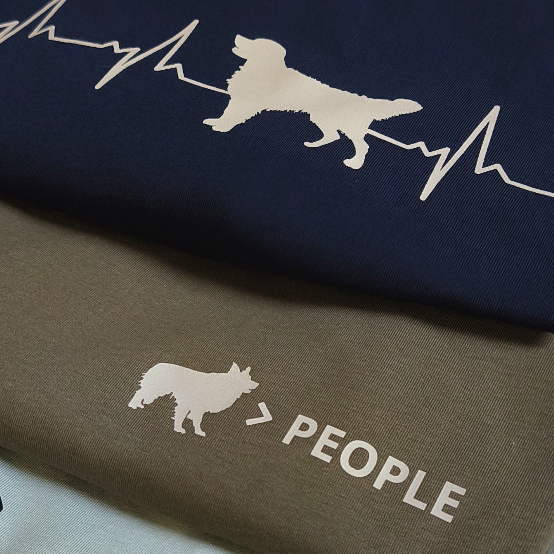 Zwei gefaltete T-Shirts in French Navy und Khaki mit Hunde-Brustprints – Herzschlagmotiv mit Hundesilhouette und „Hund > People“-Motiv.